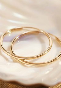 Gold hoop earrings with a smooth, shiny surface, displayed on a shell-shaped dish. The design is circular and minimalist.