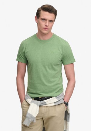 Młody mężczyzna ubrany w zielony t-shirt z krótkim rękawem, beżowe spodnie, zegarek i szaro-biały pasiasty sweter zawiązany wokół talii.