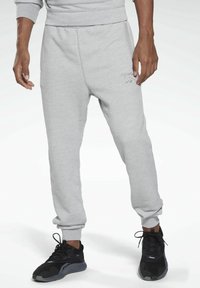 Mann trägt hellgraue Reebok-Sweatpants und schwarze Sportschuhe, steht vor einem schlichten weißen Hintergrund.