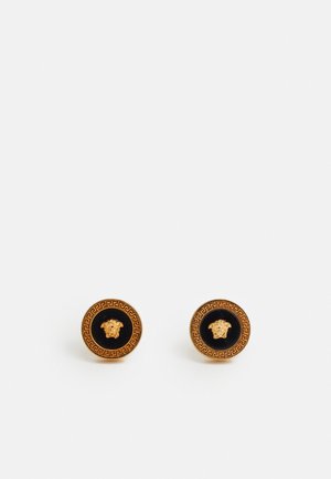 Brincos redondos com um centro preto e um detalhe de cabeça de leão dourada, rodeados por um padrão decorativo grego em ouro.