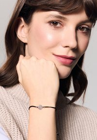 Czarna bransoletka z kamieniem szlachetnym w kształcie kwiatu, noszona na nadgarstku; modelka ma na sobie sweter w neutralnym kolorze wykonanego na drutach.