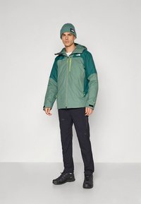 Men's two-tone jacket in green and teal with a zip front, adjustable hood, and bright yellow accents; paired with black pants and shoes.