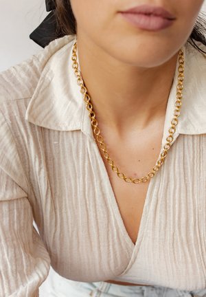 Goldkette mit runden Gliedern, kombiniert mit einer blass gerippten beigefarbenen Bluse mit spitzem Kragen.