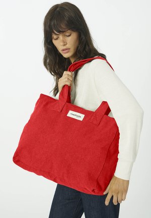 Femme aux cheveux noirs portant un top blanc à manches longues et un jean, tenant un grand sac fourre-tout rouge marqué "rivedroite" sur son épaule.