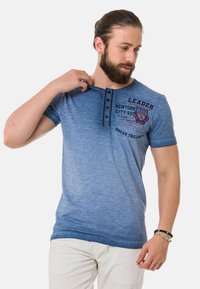 Blaues T-Shirt mit kurzen Ärmeln und einem Farbverlauf-Design, ausgestattet mit einer Knopfleiste und grafischen Details in dunklen Farben.