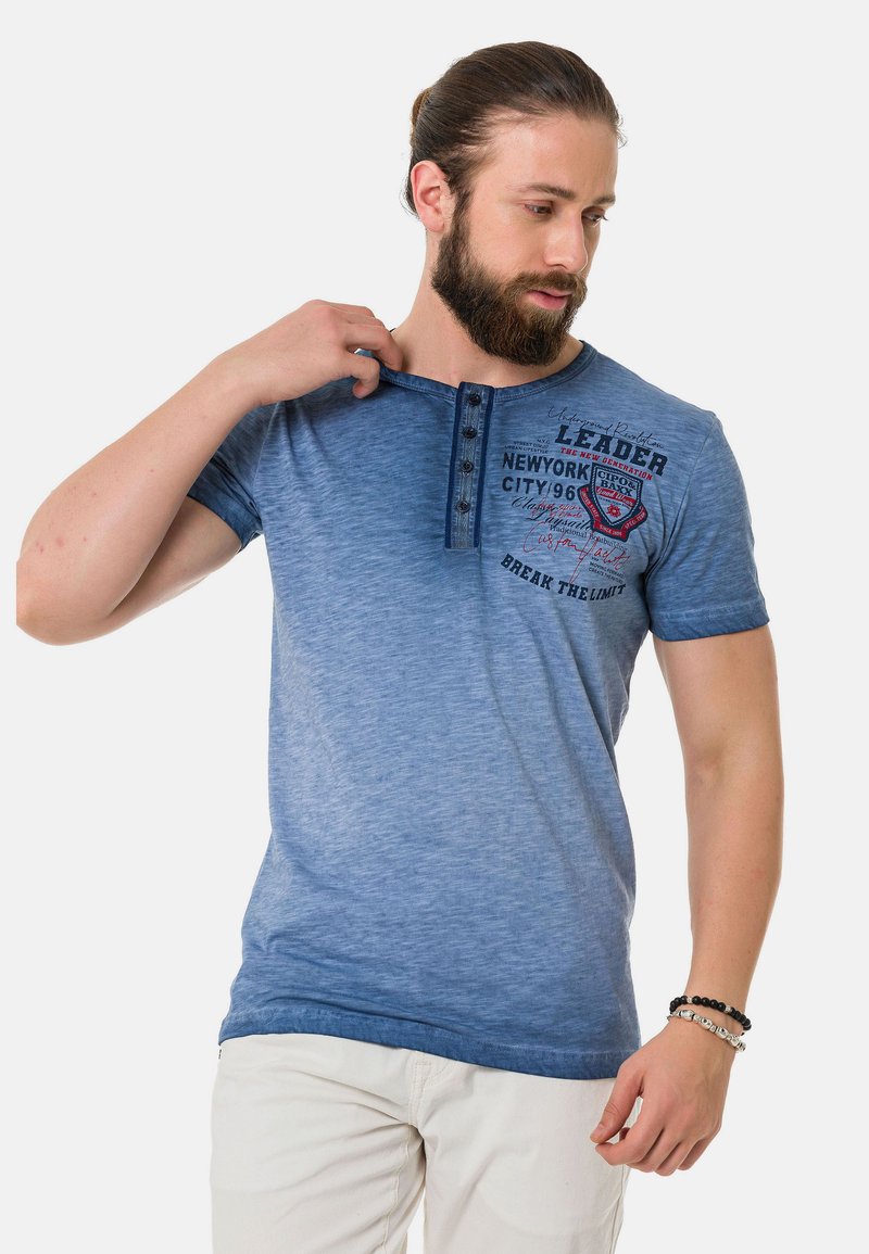 Blaues T-Shirt mit kurzen Ärmeln und einem Farbverlauf-Design, ausgestattet mit einer Knopfleiste und grafischen Details in dunklen Farben.