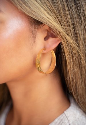 Pendientes de aro de oro con una superficie texturizada, de forma grande de media luna y un acabado pulido, colgando de una oreja perforada.