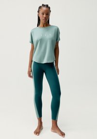 Svetlozelený top s krátkym rukávom a potlačeným logom v kombinácii s tehlovými legínami s kontrastným bočným panelom. Hladká textúra, priliehavý strih.