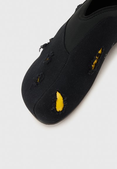 Μαύρο αθλητικό παπούτσι με σκόπιμες φθαρμένες οπές που αποκαλύπτουν κίτρινο ύφασμα από κάτω, εμφανιζόμενο σε απλό ανοιχτό φόντο.