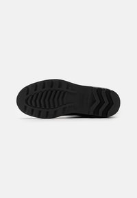 Black rubber shoe sole with a textured tread pattern, featuring zigzag grooves for grip and stability. Smooth finish with minimal branding.