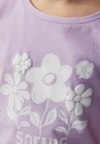 Lila T-shirt met witte bloemgraphics en textuuraccenten; bevat een glanzende glinsterende bloem en een zachte stofafwerking.