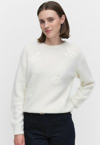 Jonge vrouw met kort bruin haar, gekleed in een witte trui met subtiele bloemenpatronen en donkere broeken, staand met één hand op haar heup.