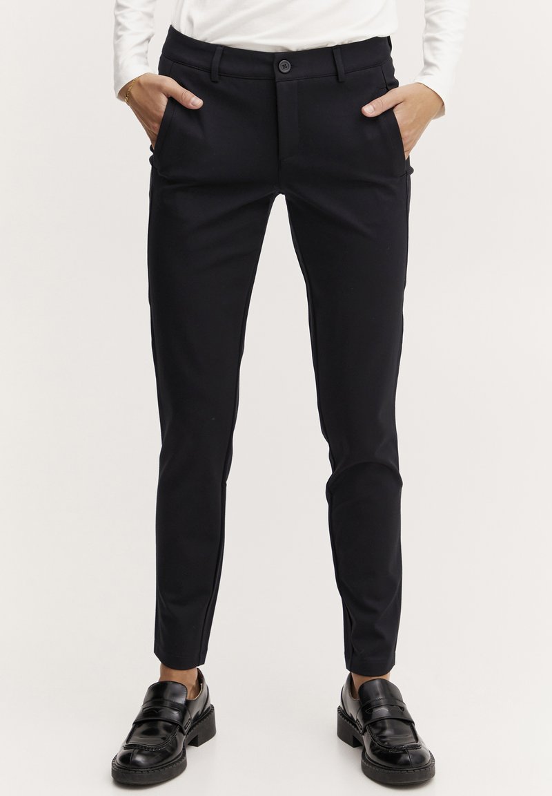 Schwarze, schmal geschnittene Hose mit glatter Textur, Knopfverschluss, Gürtelschlaufen und seitlichen Taschen. Getragen mit schwarzen Schuhen.