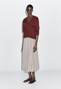 Mørkerød slåomtrøje med lange ærmer, kombineret med et lyst plisseret midinederdel. Modellen bærer sorte solbriller og brune flade sandaler.