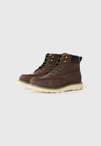 Bottes chelsea en cuir marron avec des accents noirs, des lacets verts et noirs, et une semelle en caoutchouc crème. Caractéristiques de couture et d'un motif texturé.