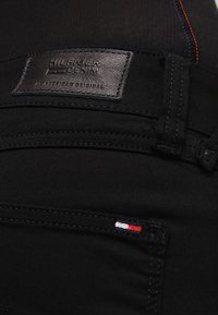 Jeans de mezclilla negra con un parche de cuero de la marca, con costuras en contraste en rojo y azul. Detalle notable de una pequeña bandera en el bolsillo.