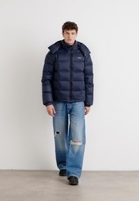 Navy puffer jacket with a zip closure and removable hood, paired with light blue distressed jeans and black shoes.