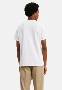 Mężczyzna stojący tyłem do kamery, ubrany w gładką białą koszulkę z krótkim rękawem i beżowe spodnie, na jednolitym tle.