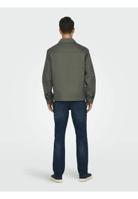 Chaqueta gris con un cuello estructurado, puños con botones y detalle en el yugo trasero, combinada con jeans de mezclilla azules y calzado informal.