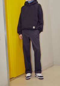 Felpa nera con cappuccio, tasca frontale, cordino regolabile e patch con logo. Abbinata a pantaloni scuri e sneakers bianche con accenti neri. Porta gialla.