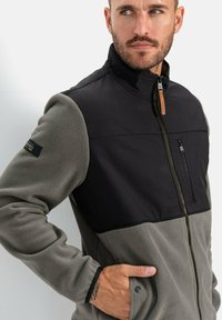Fleecejacke mit schwarzem Oberteil und grauem Unterteil. Verfügt über eine zipped Brusttasche und einen hohen Kragen. Weiche Textur und kontrastierende Nähte.