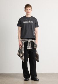 Hallhalli puuvillane t-särk, millel on "CALVIN KLEIN" logo, koos mustade laia lõikega pükstega ja seotud ruudulise flanell-särgiga. Mustad kingad.