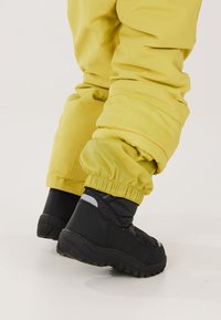 Geel gevoerde sneeuwbroek met elastische boorden, gecombineerd met zwarte waterdichte winterlaarzen met een textuur op de zolen en reflecterende accenten.