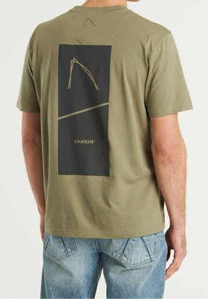 Homme portant un t-shirt vert olive avec un imprimé rectangulaire noir et le texte "CHASIN'" dans le dos, associé à un jean en denim bleu clair.