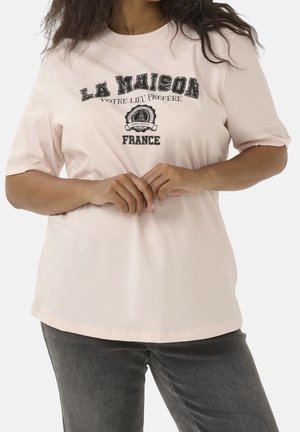 Osoba nosząca bladoróżowy t-shirt z czarnym francuskim napisem i grafiką, w połączeniu z ciemnoszarymi dżinsami, ręce splecione na wysokości talii.