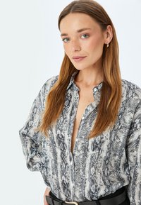 Blusa con estampado de serpiente en blanco y negro, con cuello tipo camisa, cierre de botones y mangas largas. Textura mate.