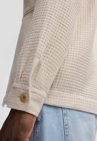 Camicia beige a coste waffle con tessuto testurizzato, dettagli con polsini abbottonati e vestibilità rilassata, abbinata a jeans in denim azzurro chiaro.
