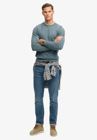 Sudadera de color teal claro con cuello redondo, logo azul, combinada con jeans azules y botas beige. Camisa de cuadros gris atada alrededor de la cintura.