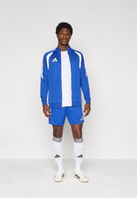 Μπλε αθλητική φόρμα με λευκές ρίγες, σετ με λευκό πουκάμισο, μπλε σορτς και ριγέ κάλτσες. Το μοντέλο φοράει ποδοσφαιρικά παπούτσια.