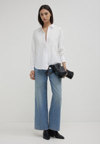 Weißes Button-up Hemd mit einer vorderen Naht, langen Ärmeln. Kombiniert mit hellblauen weit geschnittenen Jeans und schwarzen Stiefeletten, hält eine schwarze Tasche.