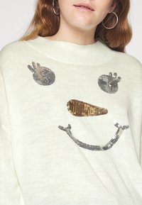 Weißer Strickpullover mit einem Pailletten-Schneemann-Gesicht, silbernen Augen, einer goldenen Karotten-Nase und einem silbernen Pailletten-Lächeln.