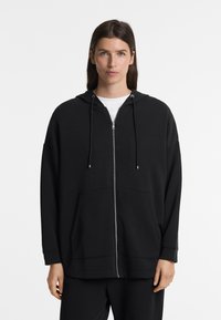 Crna zip-up hoodie s opuštenim krojem, velikim prednjim džepovima, podesivim vezicama na kapuljači i mekanom teksturom materijala. Dugi rukavi.