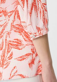 Bluse mit rotem und weißem Blumenmuster, Puffärmeln und einem Knopfakzent an den Bündchen; hergestellt aus einem leichten, glatten Stoff.