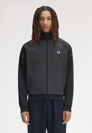 Junge Person mit langen Dreadlocks, die eine schwarze Jacke mit Reißverschluss und kleinem weißem Lorbeer-Logo auf der Brust trägt.