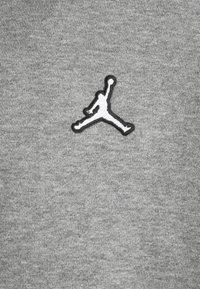 Grå sweatshirt-tyg med en broderad svart och vit Jumpman-logga på vänster bröst. Mjuk textur med en slät yta.