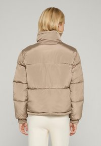 Jachetă bej căptușită cu guler înalt, având un finisaj satinat neted și manșete elastice. Designul matlasat adaugă textură și dimensiune.