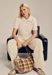 Polo crème à texture shaggy, pantalon beige à jambes larges, sac hobo à carreaux marron à motif, baskets grises et dorées, assis sur une chaise noire.