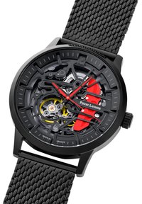 Reloj de estilo esqueleto con caja negra, correa texturizada negra, acentos rojos y mecánica interna visible. Presenta marcadores de hora y manecillas audaces.