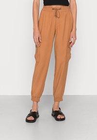 Pantalones cargo en un color marrón cálido, con una banda elástica en la cintura y cordón, tobillos ajustados y bolsillos laterales. Combinados con sandalias negras.