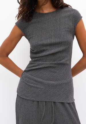 Femme portant un haut gris côtelé ajusté sans manches et un pantalon gris côtelé assorti sur un fond blanc uni.