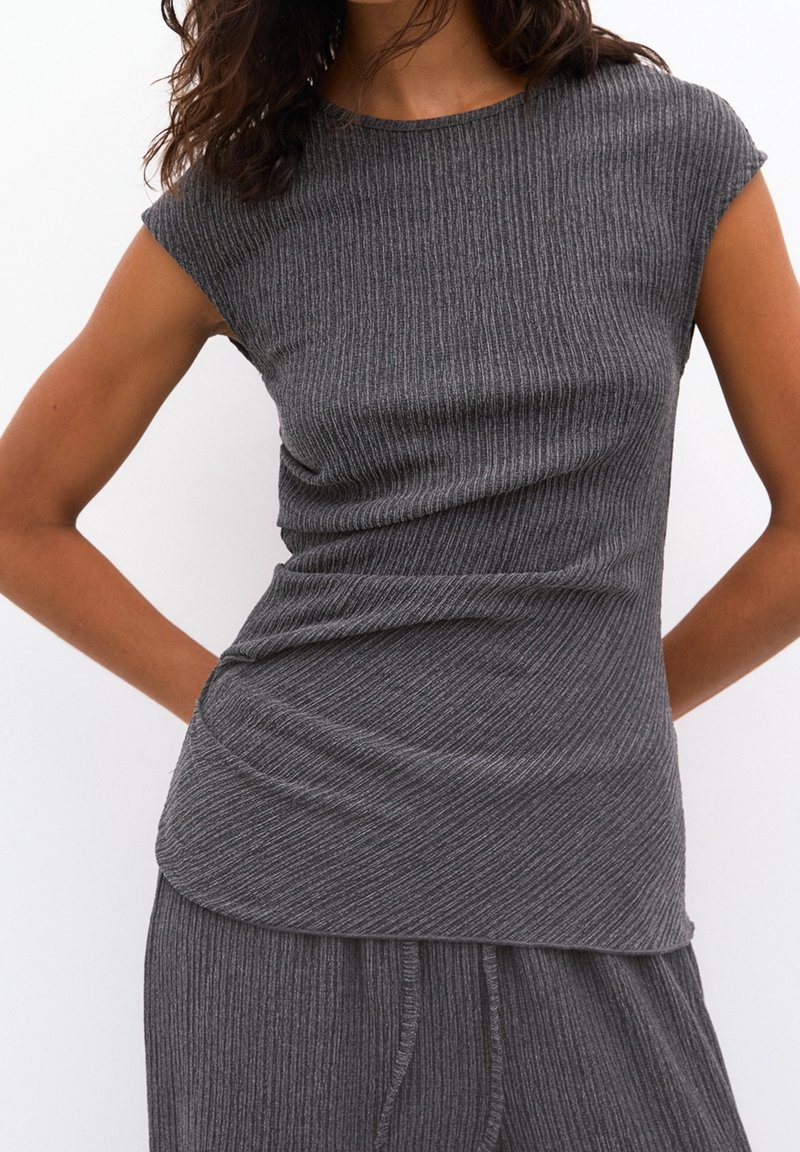 Femme portant un haut gris côtelé ajusté sans manches et un pantalon gris côtelé assorti sur un fond blanc uni.