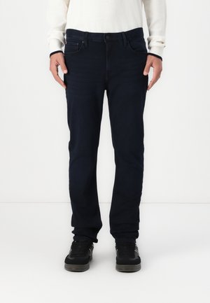 Mann trägt dunkelblaue Straight-Leg-Jeans, weißes langärmeliges Hemd und schwarze Sneakers und steht vor einem schlichten weißen Hintergrund.