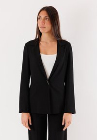 Blazer nero su misura con rever a punta, chiusura con un solo bottone, maniche lunghe e una texture liscia, abbinato a una maglietta bianca.