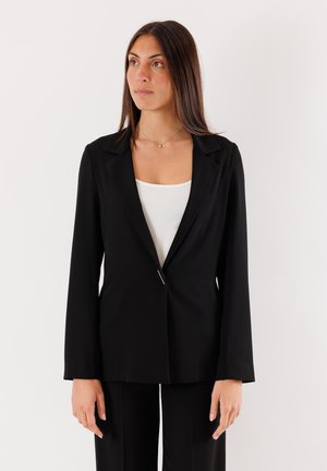 Blazer nero su misura con rever a punta, chiusura con un solo bottone, maniche lunghe e una texture liscia, abbinato a una maglietta bianca.
