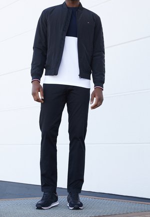 Mann trägt eine schwarze Bomberjacke über einem navy-weiß farblich abgesetzten Shirt, schwarze Hose und schwarze Sneakers, steht auf einem Teppich im Freien.