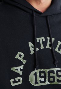 Sudadera negra que presenta una gran impresión de texto verde desgastado que dice "GAP ATH." y "1969." Incluye un cordón y tejido texturizado.